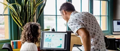 Two people look at a computer monitor displaying graphs and data in an office with large windows and a plant nearby.