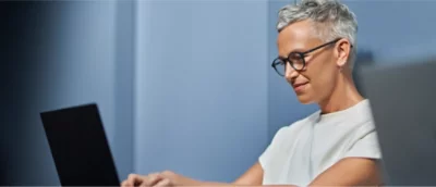 A person with short gray hair and glasses is typing on a laptop, seated in a room with blue walls.